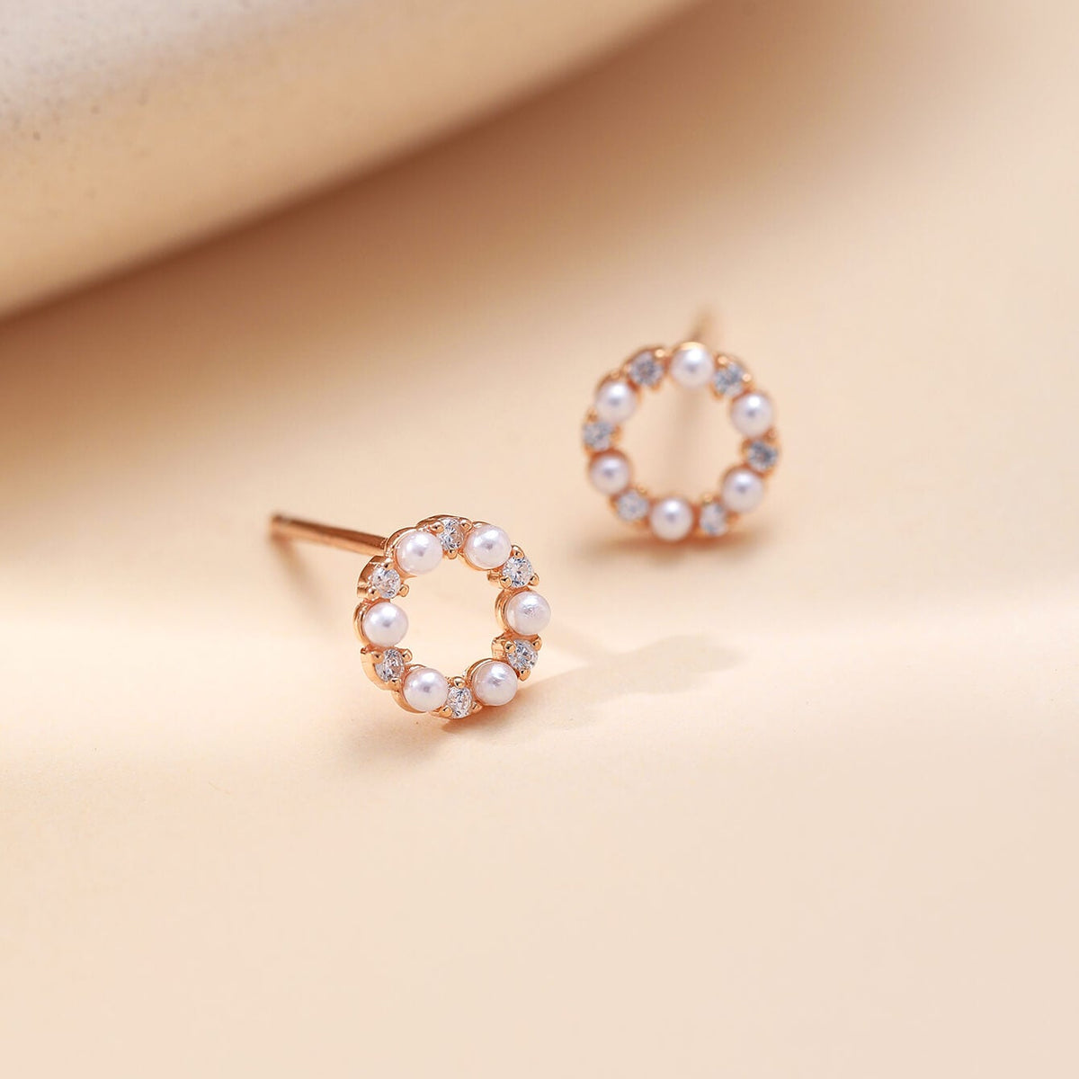 A pair of rose gold stud earrings with
small white pearls and clear crystals, displayed on a warm beige surface.