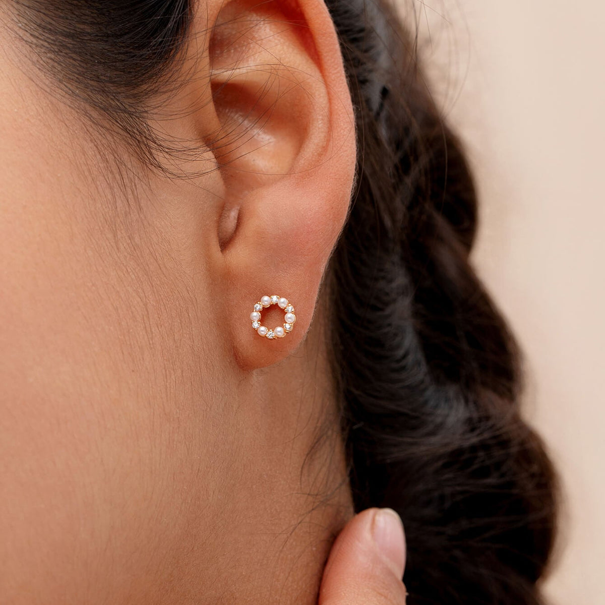 A close-up of a model's ear wearing a small stud earring with a halo of pearls and CZ stones in a warm metallic setting.