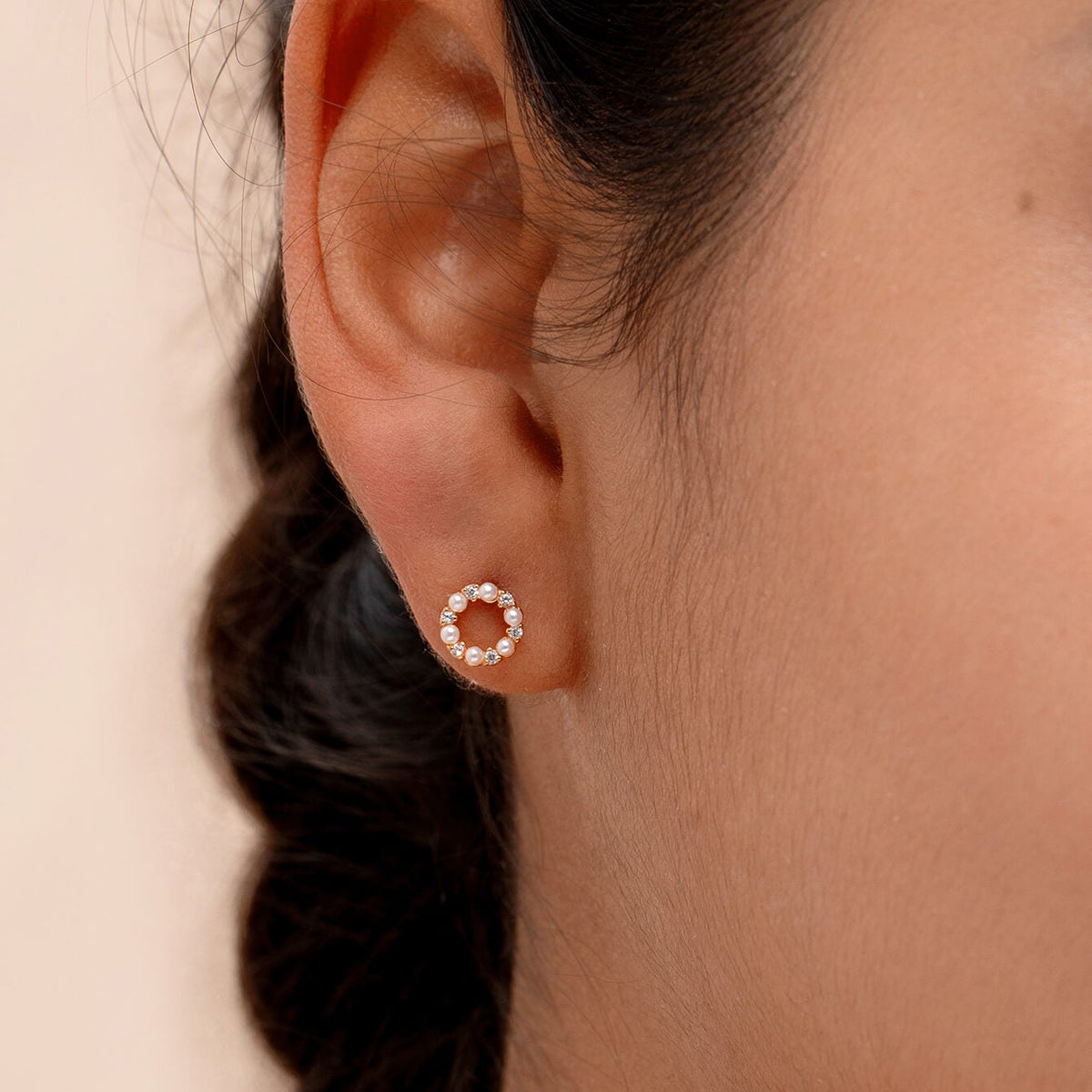 A close-up of a woman's ear wearing a small circular stud earring with tiny clear stones set in a warm metallic setting, against a soft beige background.