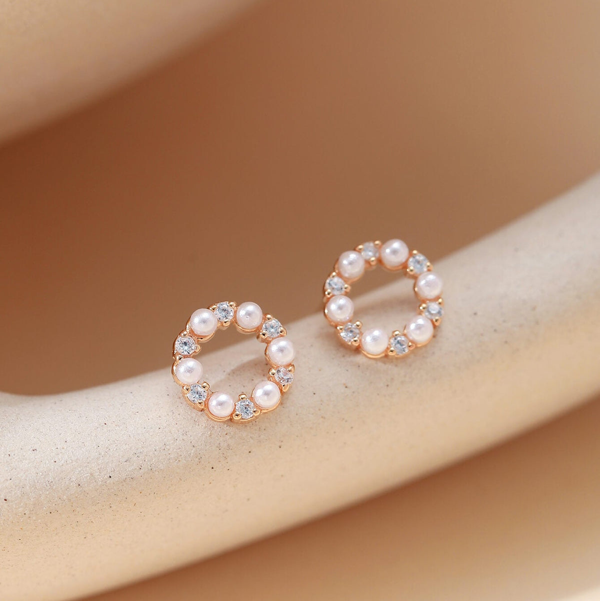 A pair of rose gold hoop-style earrings with small white pearls and clear rhinestones, displayed on a beige curved jewelry stand.