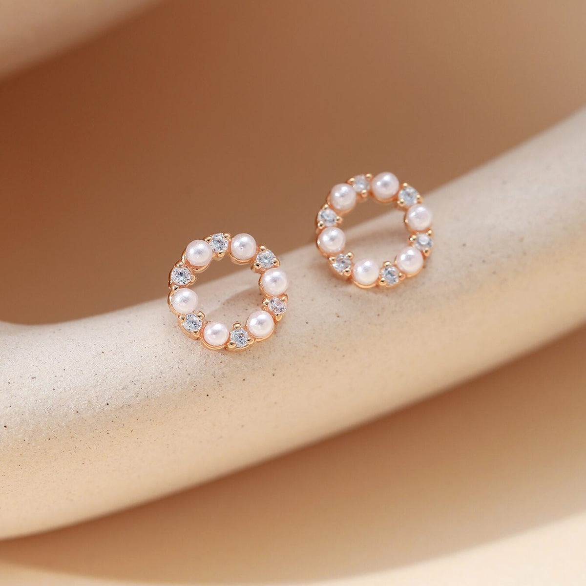 A pair of rose gold hoop-style earrings with small white pearls and clear rhinestones, displayed on a beige curved jewelry stand.