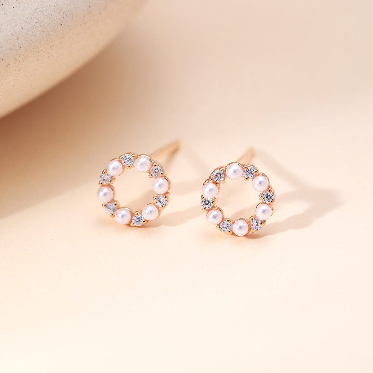 A pair of rose gold hoop-style stud earrings with a circular arrangement of white faux pearls and small clear rhinestones, displayed on a warm beige surface.