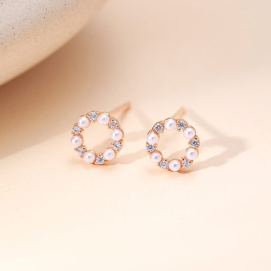 A pair of rose gold hoop-style stud earrings with a circular arrangement of white faux pearls and small clear rhinestones, displayed on a warm beige surface.