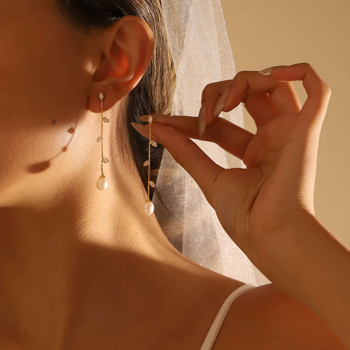 A close-up lifestyle shot of a woman wearing delicate gold dangle earrings with white freshwater pearls and small clear stones, with one earring hanging on her ear and the other being adjusted on her finger.
