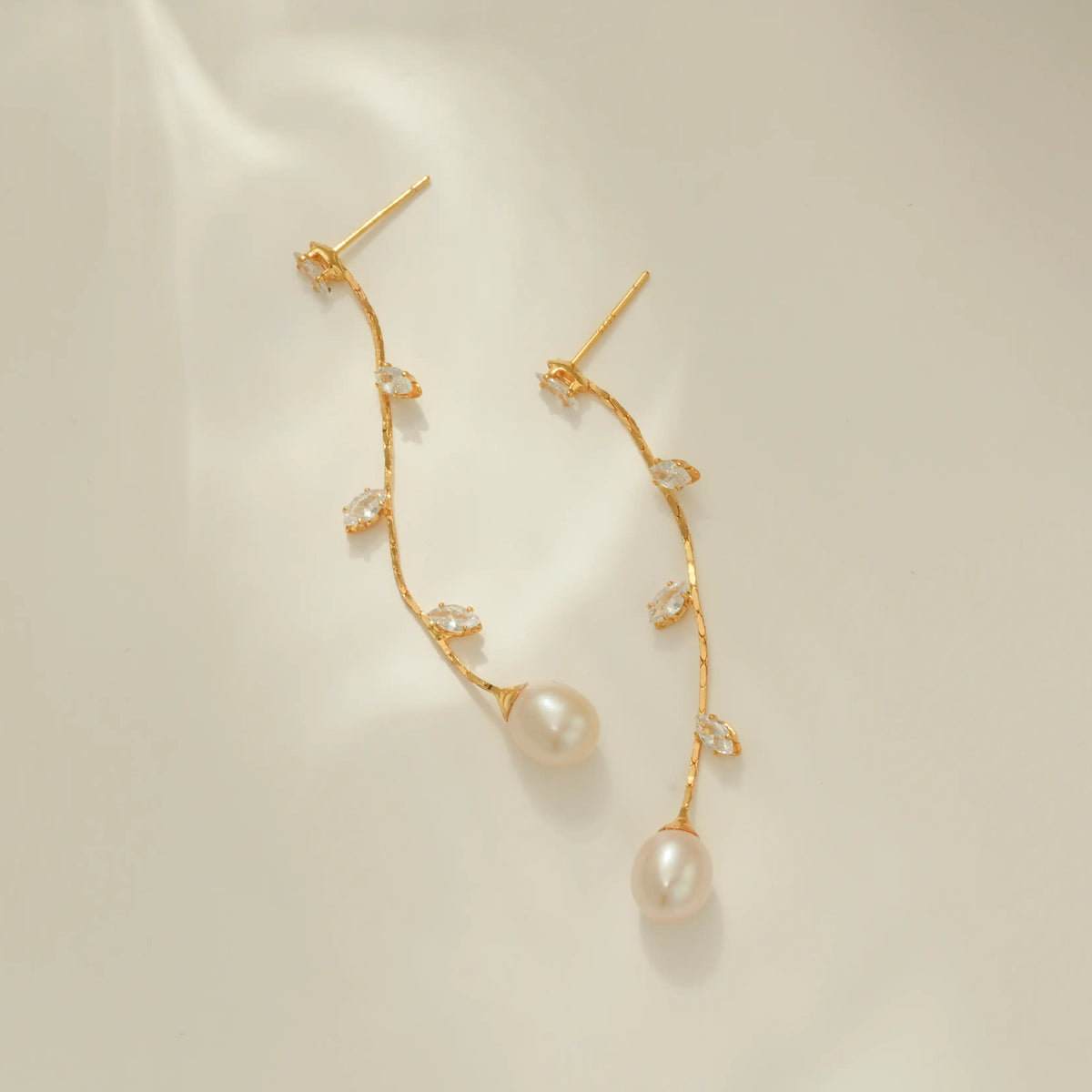 A pair of gold dangle earrings with small clear CZ leaves and a large pearl drop at the end, displayed on a clean off-white surface.