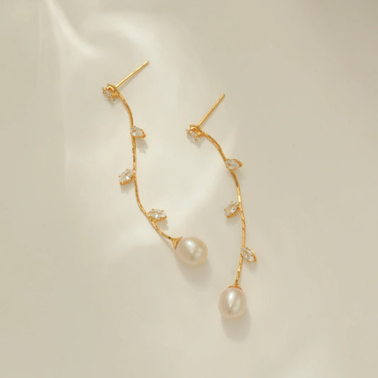 A pair of gold dangle earrings with small clear CZ leaves and a large pearl drop at the end, displayed on a clean off-white surface.