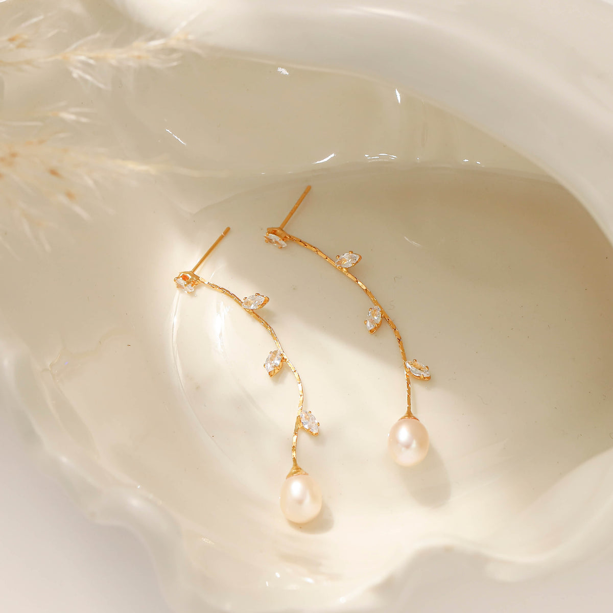 A pair of gold drop earrings with small clear crystal leaves and a teardrop-shaped pearl dangle, displayed inside a white clam shell.