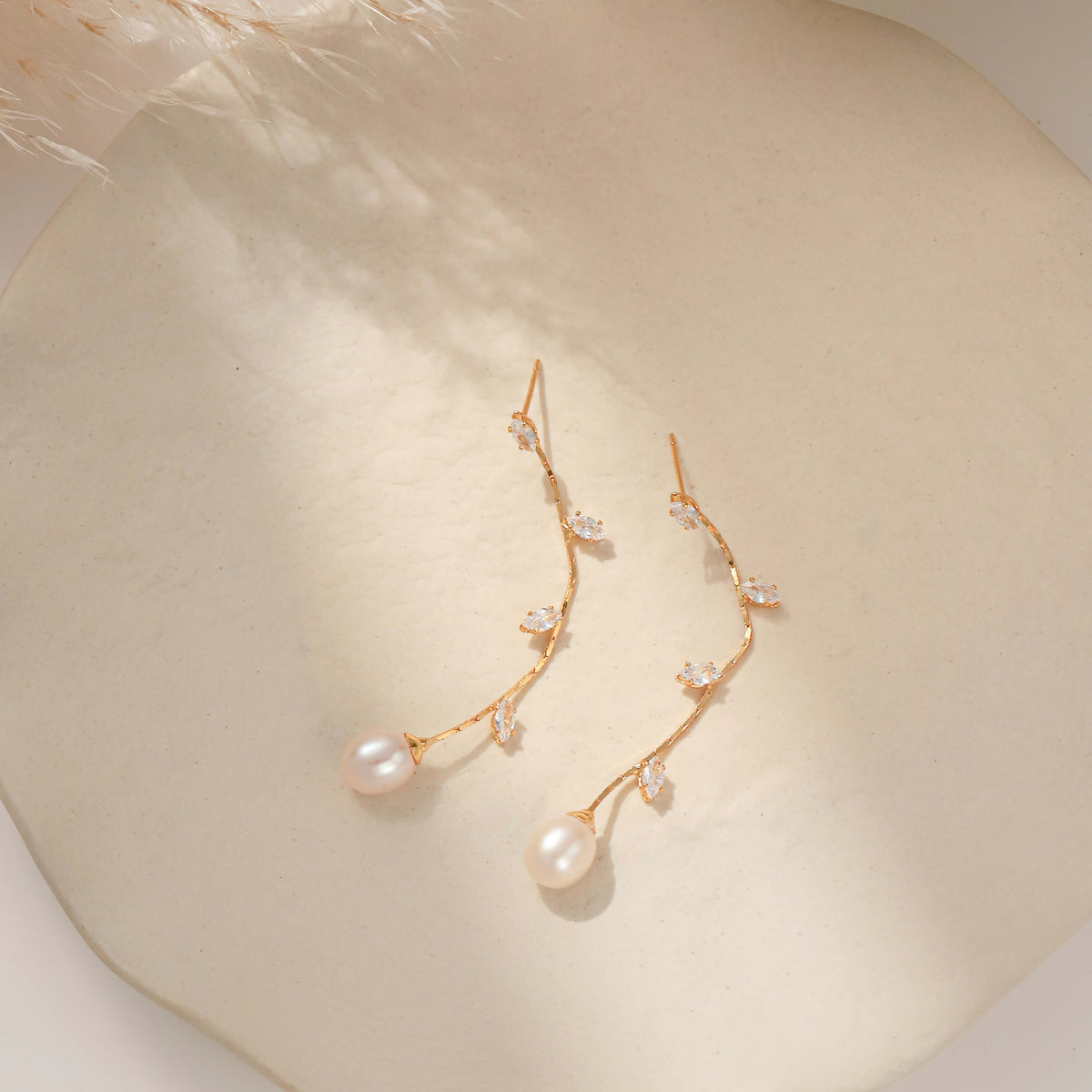 A pair of gold drop earrings with white pearl and tiny cz leaves, displayed on a textured beige surface with soft decorative grass in the corner.