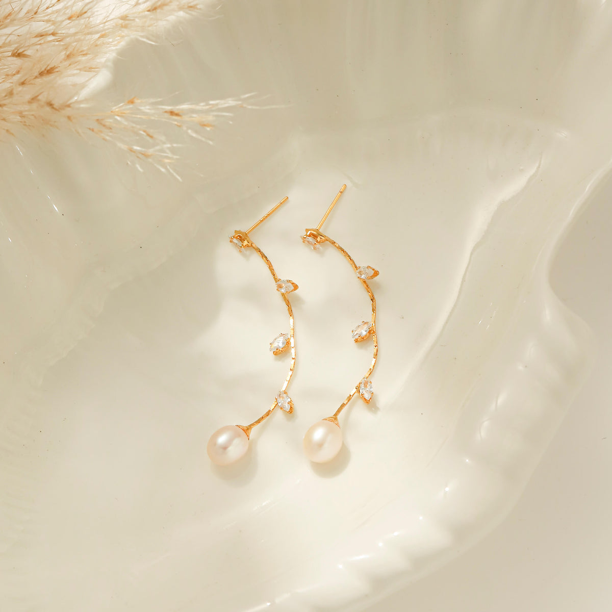 A pair of gold vine-style drop earrings with small clear stones and white pearl drops, displayed on a glossy white ceramic dish.