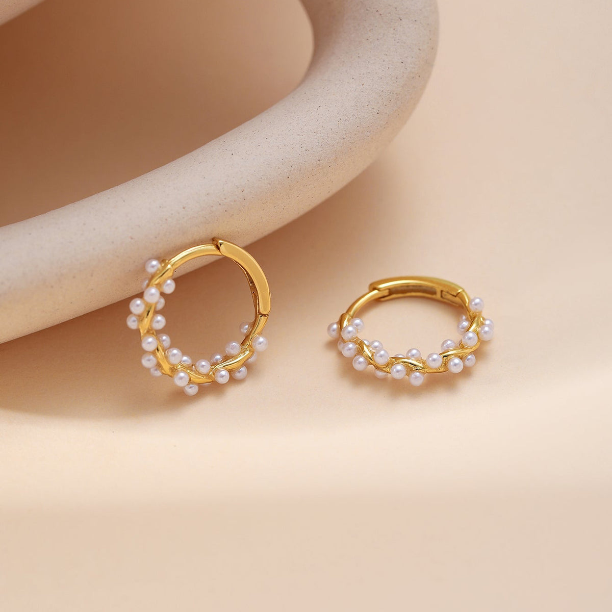 A pair of gold huggie twist hoop earrings with small white pearls, displayed on a warm beige background with a curved beige prop.