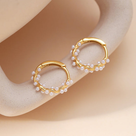 A pair of gold twisted hoop pearls earrings decorated with small white pearls, displayed on a beige textured jewelry stand.