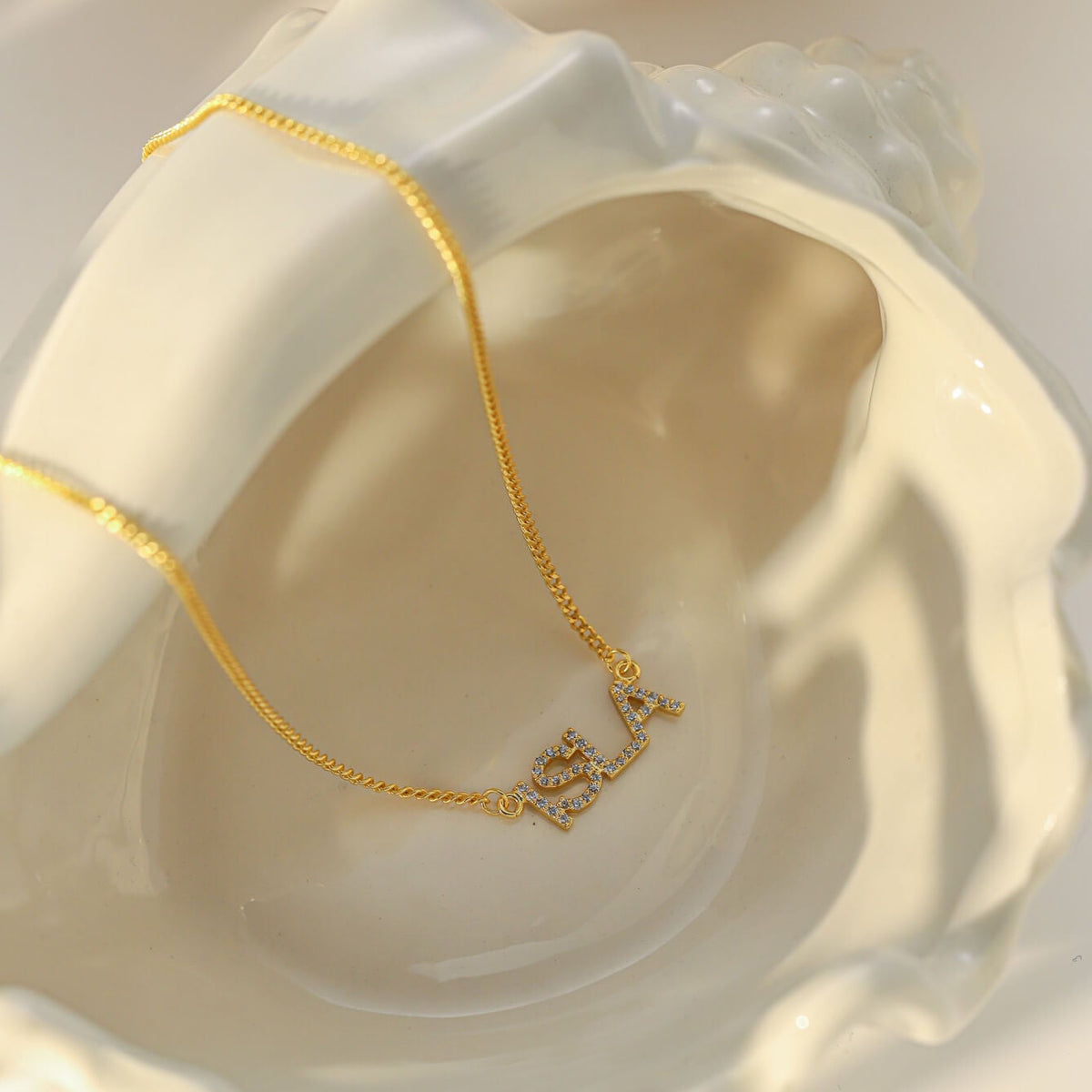 A delicate gold chain necklace with a small script nameplate pendant, displayed inside a white ceramic shell dish.