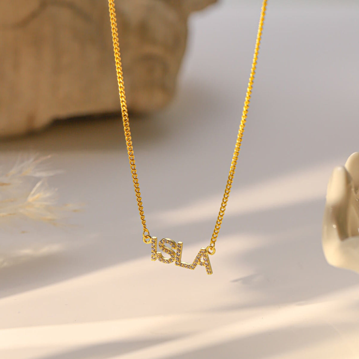 A gold chain personalised necklace with a cursive 'ISLA' pendant, displayed on a light beige surface with soft shadows and a blurred decorative stone in the background.