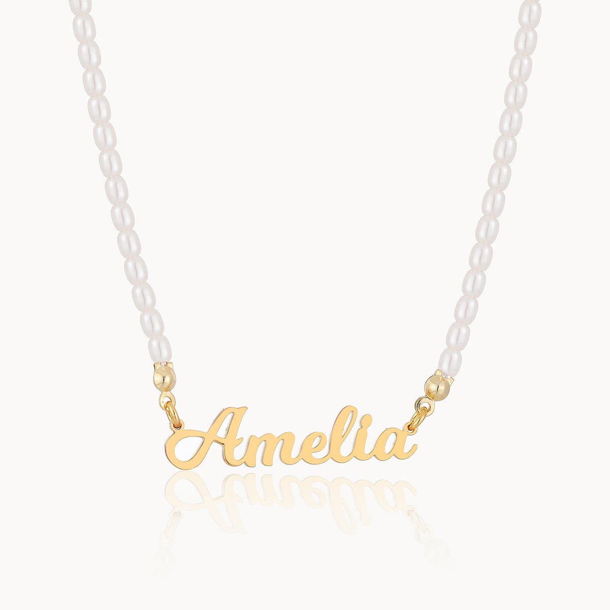 A personalized name necklace featuring a gold a white pearl chain, displayed on a clean off-white background with a soft reflection below.