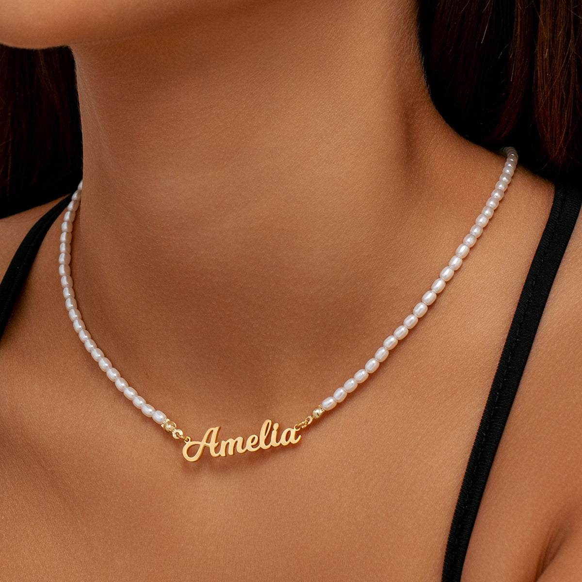 A close-up of a model's neck and chest wearing a delicate white pearl necklace with a gold nameplate spelling 'Amelia'.