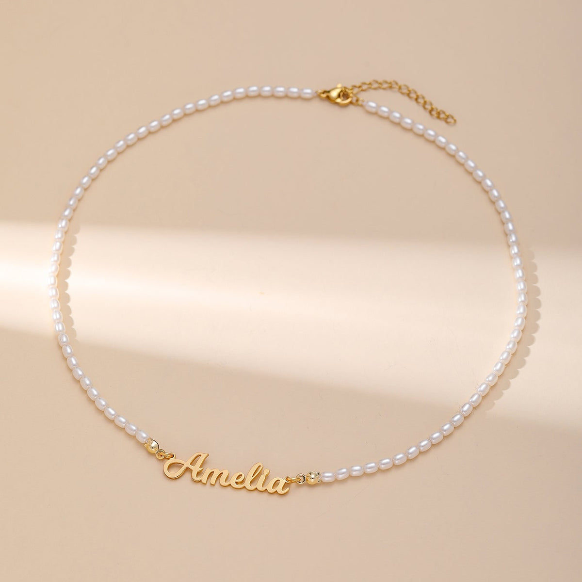A white pearl necklace with a gold custom reading 'Amelia' in cursive, displayed on a warm beige background.