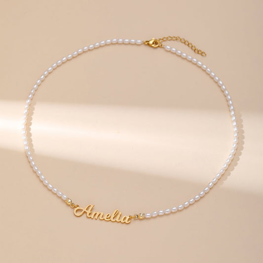 A white pearl necklace with a gold custom reading 'Amelia' in cursive, displayed on a warm beige background.