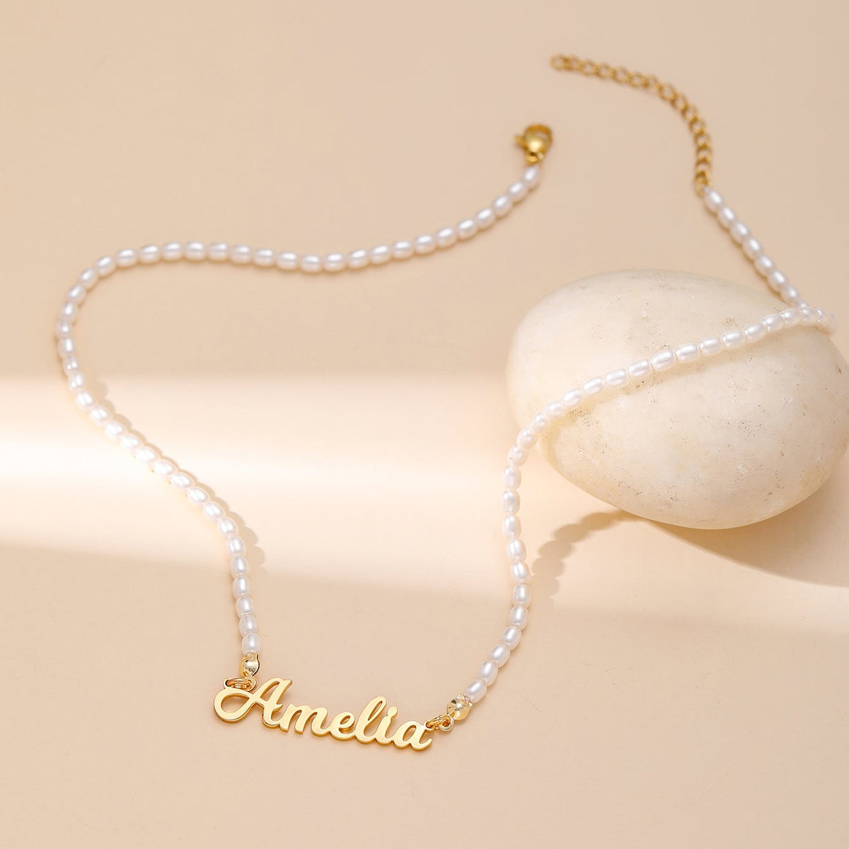 A delicate necklace with small white pearls and a gold script nameplate reading 'Amelia', styled on a warm beige background with a smooth white stone prop.