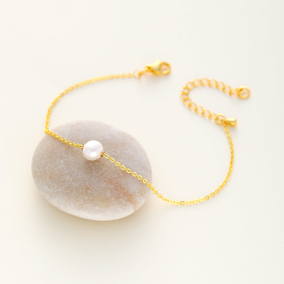 A delicate gold chain bracelet with a single white pearl bead, displayed on a smooth, light beige stone against a warm off-white background.