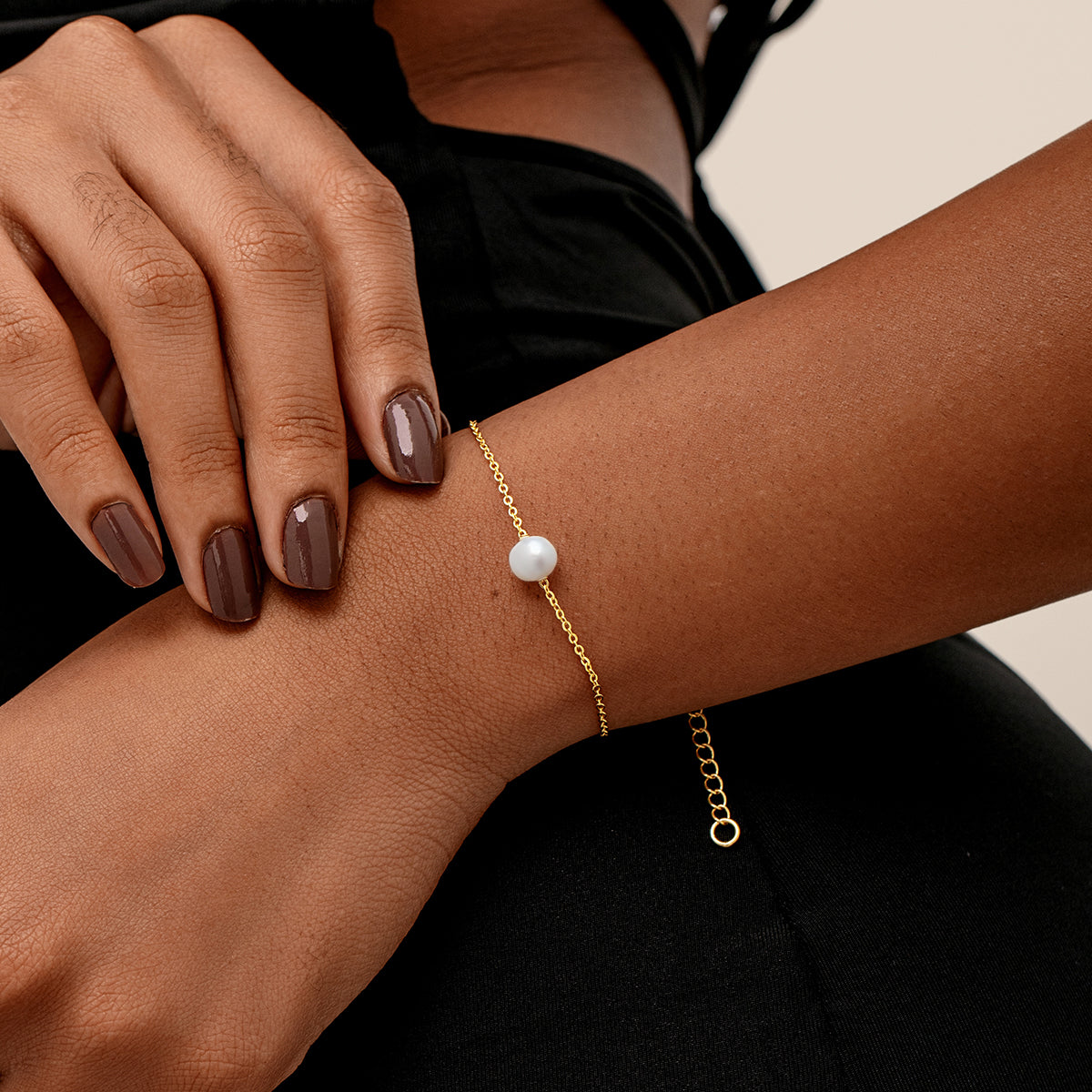 Gold minimalist single pearl bracelet, shown worn on a person's wrist. The person is wearing a black top, and their manicured hand rests near the wrist.