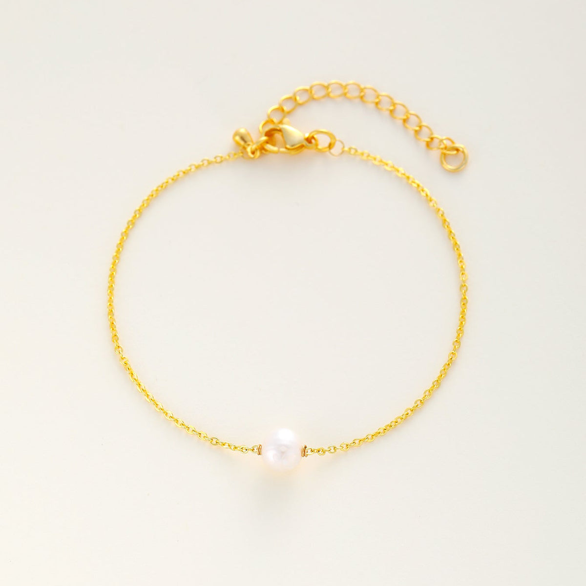 A delicate dainty gold bracelet featuring a single white pearl bead at the center, displayed on a clean, light off-white background.