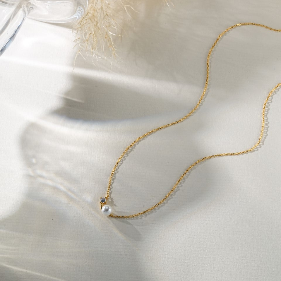 A delicate gold necklace with a small white pearl pendant and a tiny clear stone accent