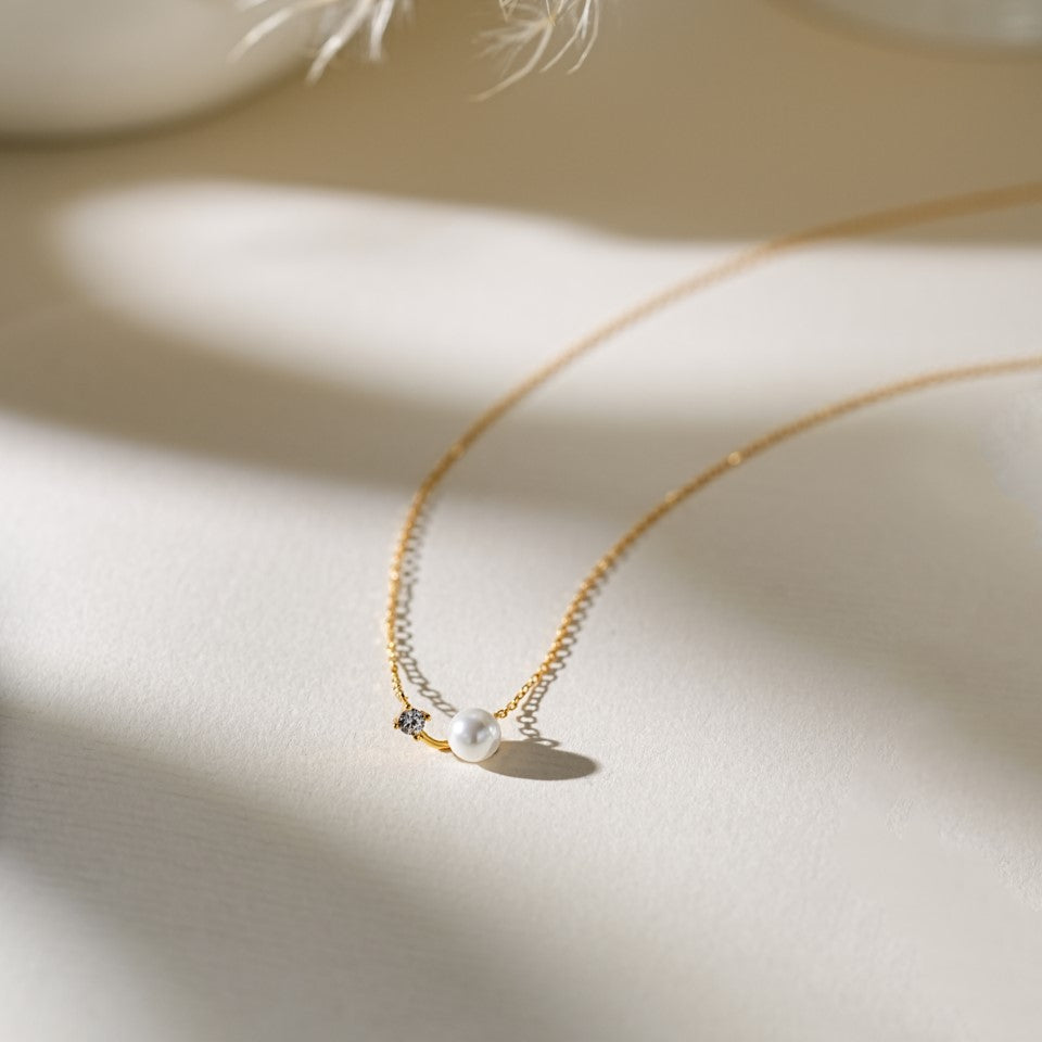 A delicate gold chain necklace with a small white pearl pendant and a tiny clear stone accent, styled on a warm beige surface with soft shadows.