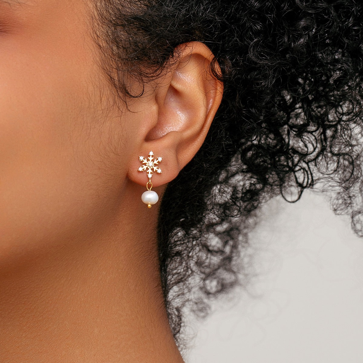 A close-up side profile of a model wearing a gold snowflake stud earring with a dangling pearl