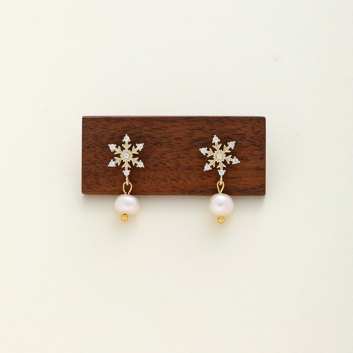 A pair of gold snowflake drop earrings with clear cz and white pearl dangles, displayed on a small dark wooden block against a light cream background.