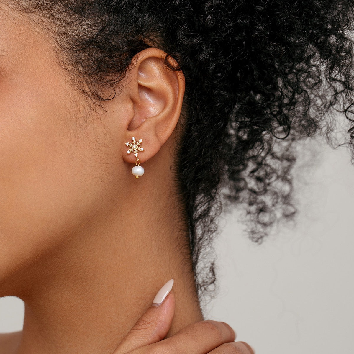A close-up of a model's ear wearing a gold snowflake stud earring with a dangling white pearl, set against a neutral light gray background.
