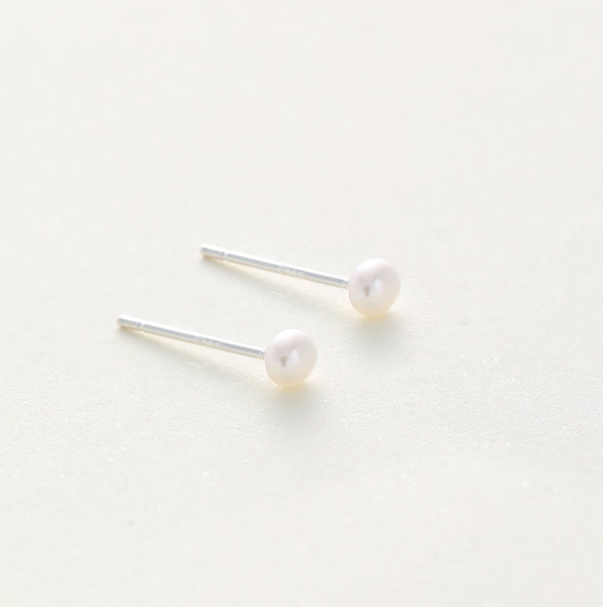 A pair of minimalist stud earrings featuring small, round white pearls on thin silver posts, displayed on a clean, light off-white background.