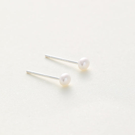 A pair of minimalist stud earrings featuring small, round white pearls on thin silver posts, displayed on a clean, light off-white background.
