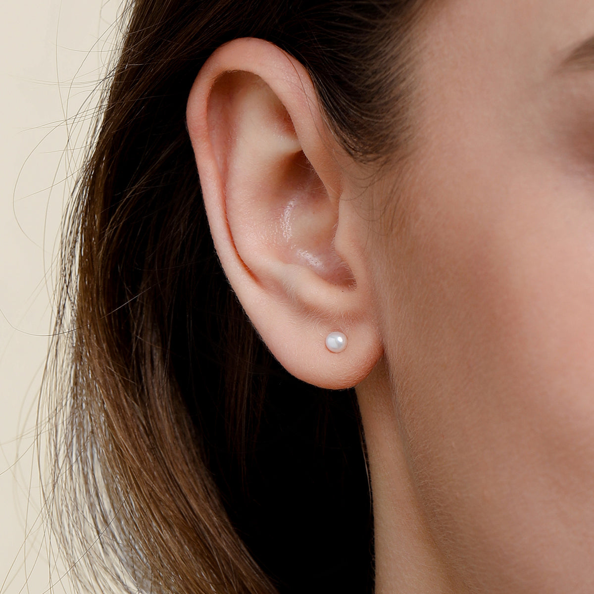 A close-up of a person's ear wearing a dainty tiny, round white pearl stud earring against a light beige background.