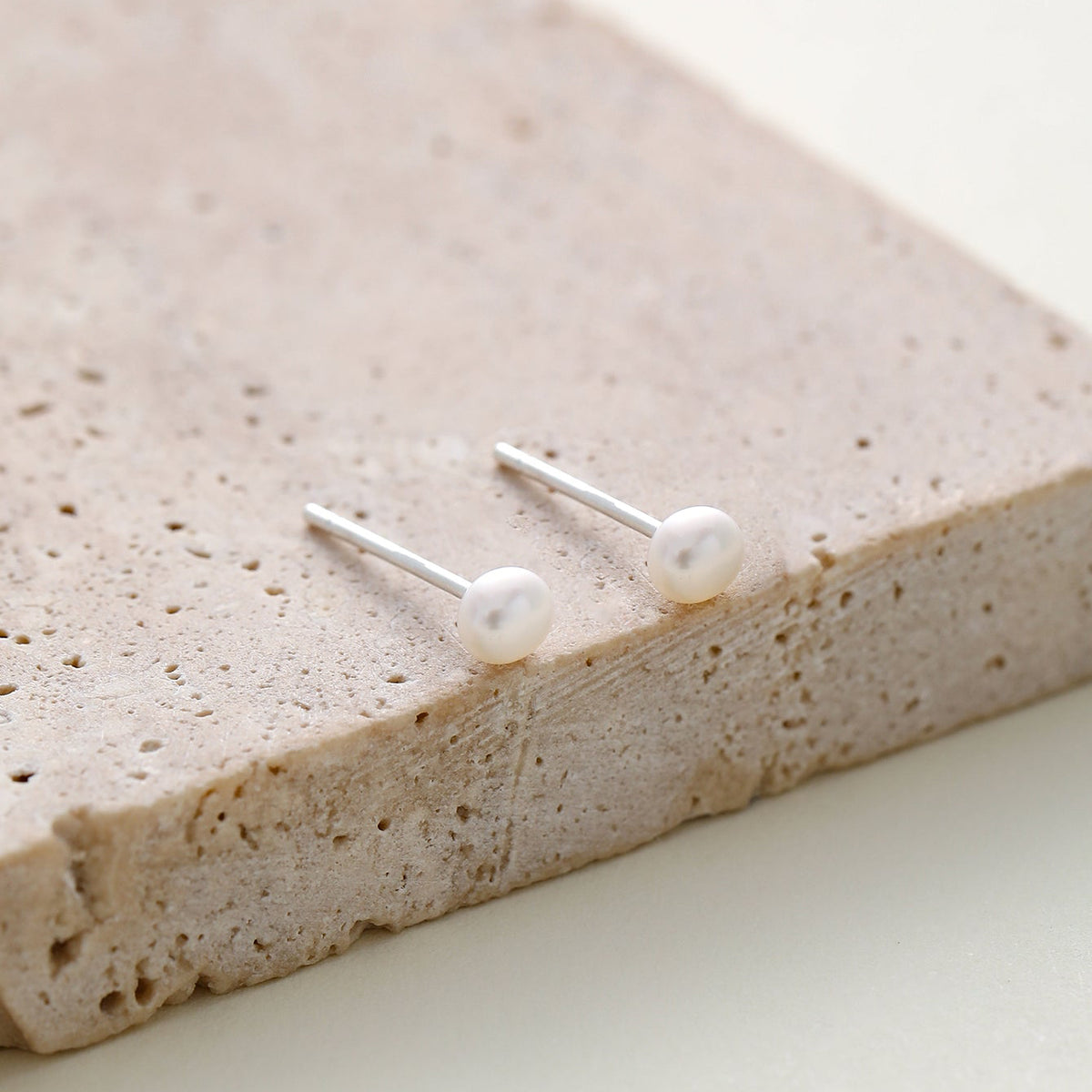 A pair of minimalist silver stud earrings with white pearl round studs, resting on a textured beige stone slab against a light cream background.