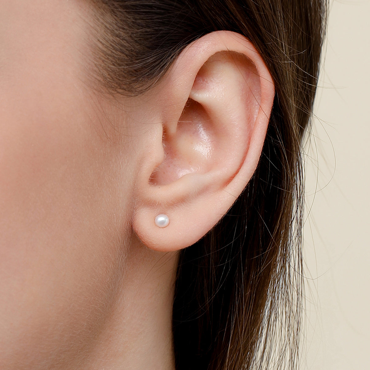 A close-up of a model's ear wearing a single small white pearl stud earring against a soft beige background.