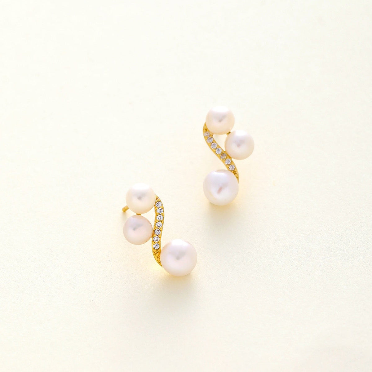 A pair of gold spiral stud earrings featuring three white freshwater pearls and a row of small clear crystals, displayed on a light cream background.