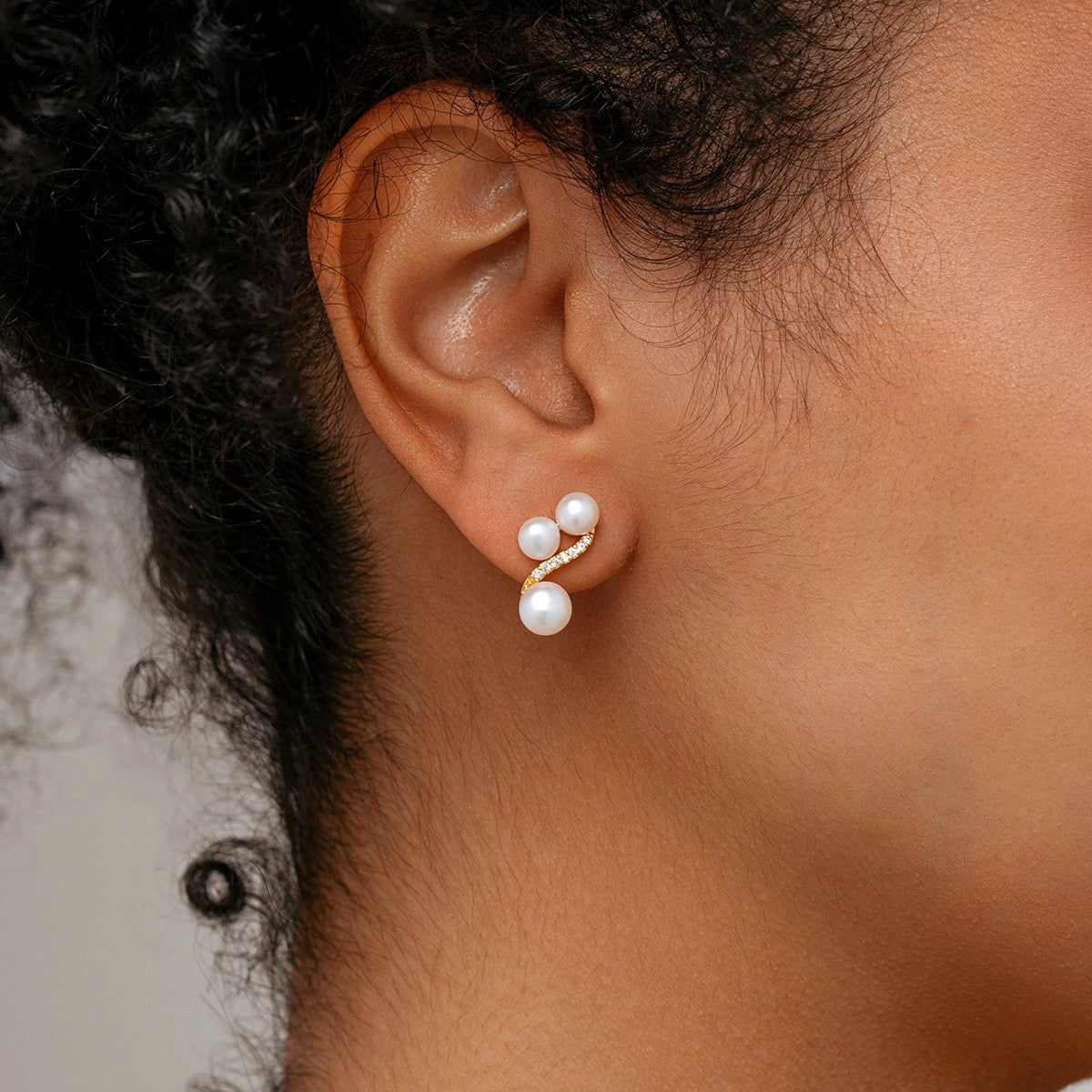A close-up of a model’s ear wearing a small triple pearls stud earrings with tiny clear stones.