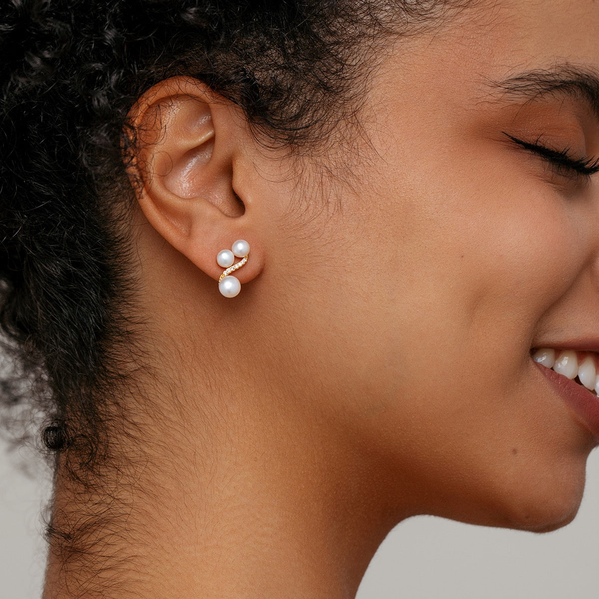 A close-up side profile of a woman wearing a small gold stud earring with three white pearls  on a light gray background.